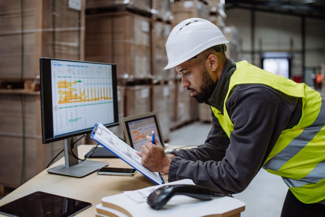 Man wearing a hard hat reviewing documents and charts in a warehouse