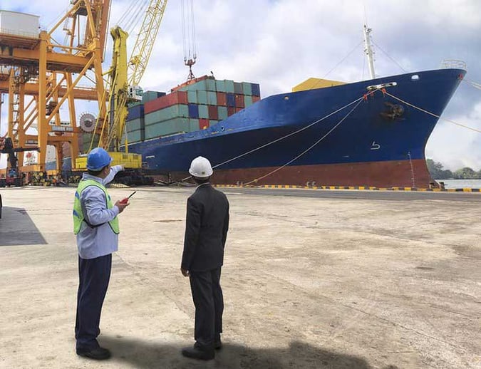 Two people with hard hats look at a cargo ship with containers