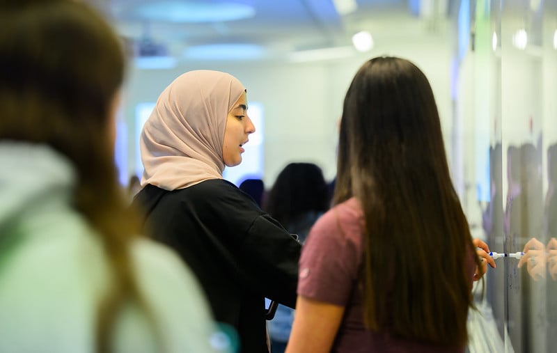 female student wearing a hijab and writing on a dry erase board with another female student listening