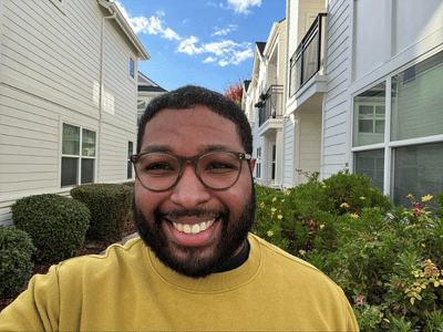 Photo of a smiling man with glasses standing outside in front of flower bushes and houses.
