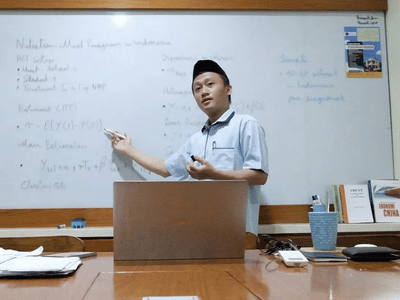 Munip Utama stands before a whiteboard, gesturing. A laptop sits open on the table in front of him.