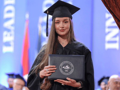 Woman with long brown hair wears a cap and gown and looks at the camera while holding her diploma