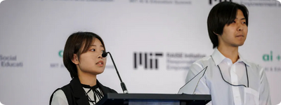 Two people stand on stage presenting their work. In the background is a white board featuring the MIT RAISE and ai + edu logos.
