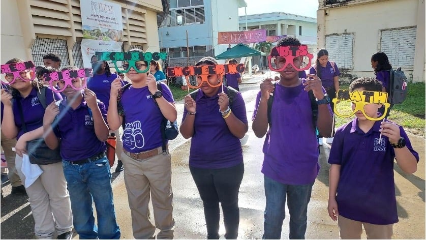 6 students wearing matching uniforms hold colorful glasses labeled with STEAM subjects, Science, Technology, Engineering, Arts and Math in front of a school building