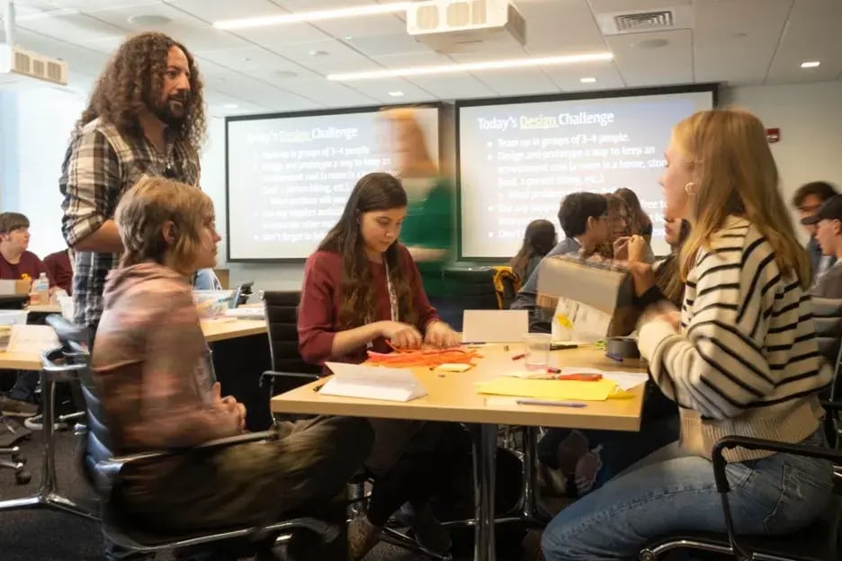 Four people sitting at a table in a classroom talk to a person standing up.