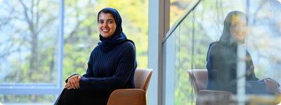 Sarah Alnegheimish sits in front of a wall of windows overlooking out-of-focus trees.