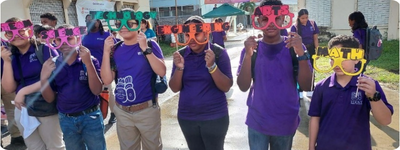 6 students wearing matching uniforms hold colorful glasses labeled with STEAM subjects, Science, Technology, Engineering, Arts, and Math in front of a school building