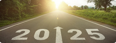The numbers “2025” with an arrow pointing straight are written in white in the middle of a road at sunset. Trees with green leaves are visible on either side of the road.