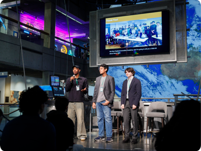 Three students stand on a stage with a large monitor behind them displaying a group of people and the words 