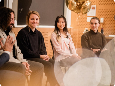 Joe Diaz, Rachael Harkavy, Joyce Yuan, Lancelot Blanchard, and Grace Song are all seated in a row in front of an audience, smiling and listening intently to someone out of the frame.