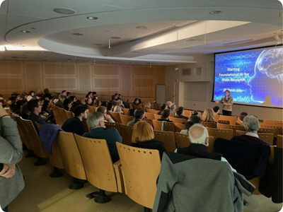 A woman speaks to a packed room of people. A large screen behind her displays a blue slide with the words, 