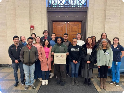 A group of people stand in front of a door. One person holds a banner that says “MICRO.